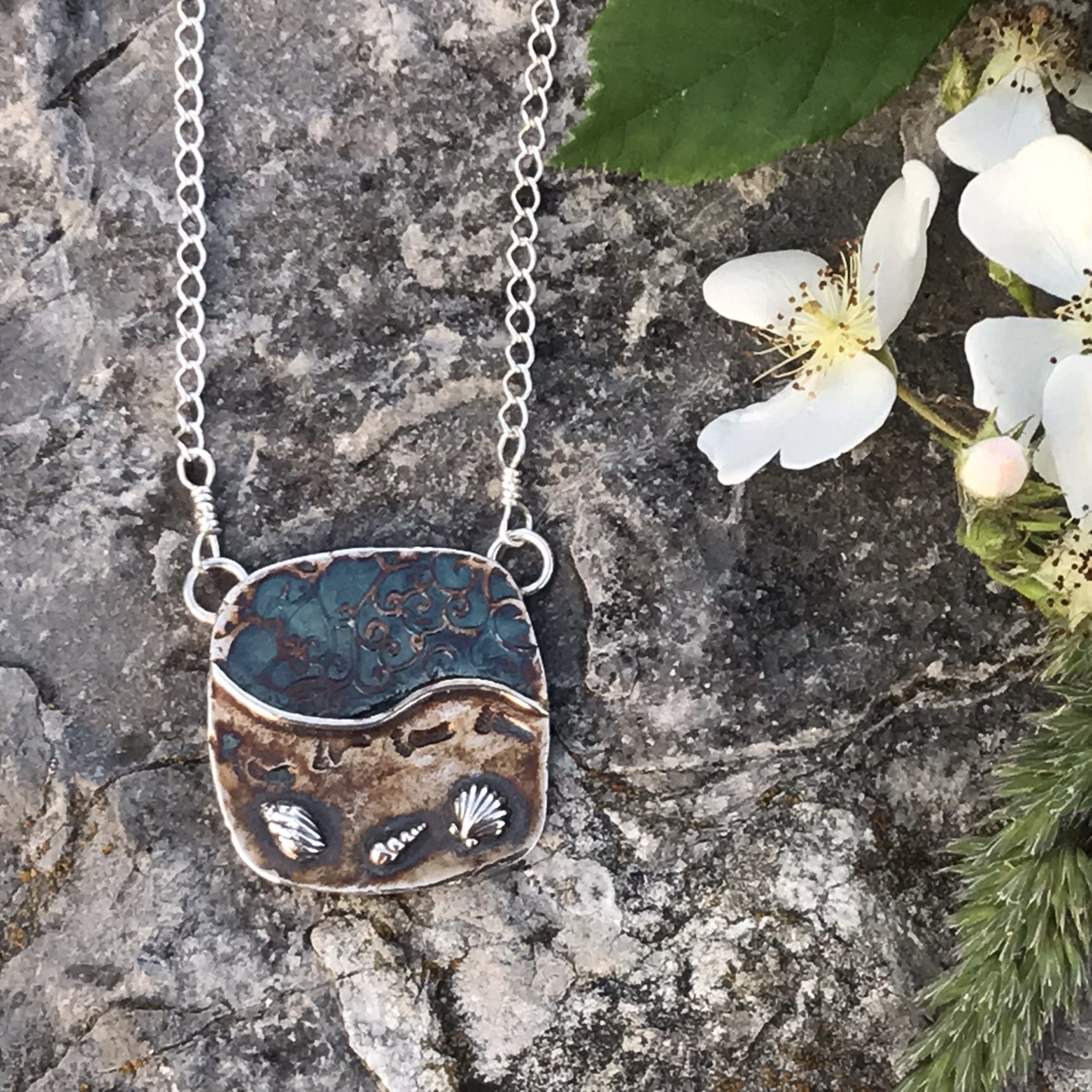 Never Alone Large Silver Cushion Pendant- 1 and 1/8 inch rounded square pendant created in fine silver metal clay and showing images of sand, waves and shells and imprinted with footprints with 18 inch sterling silver chain laying on rock