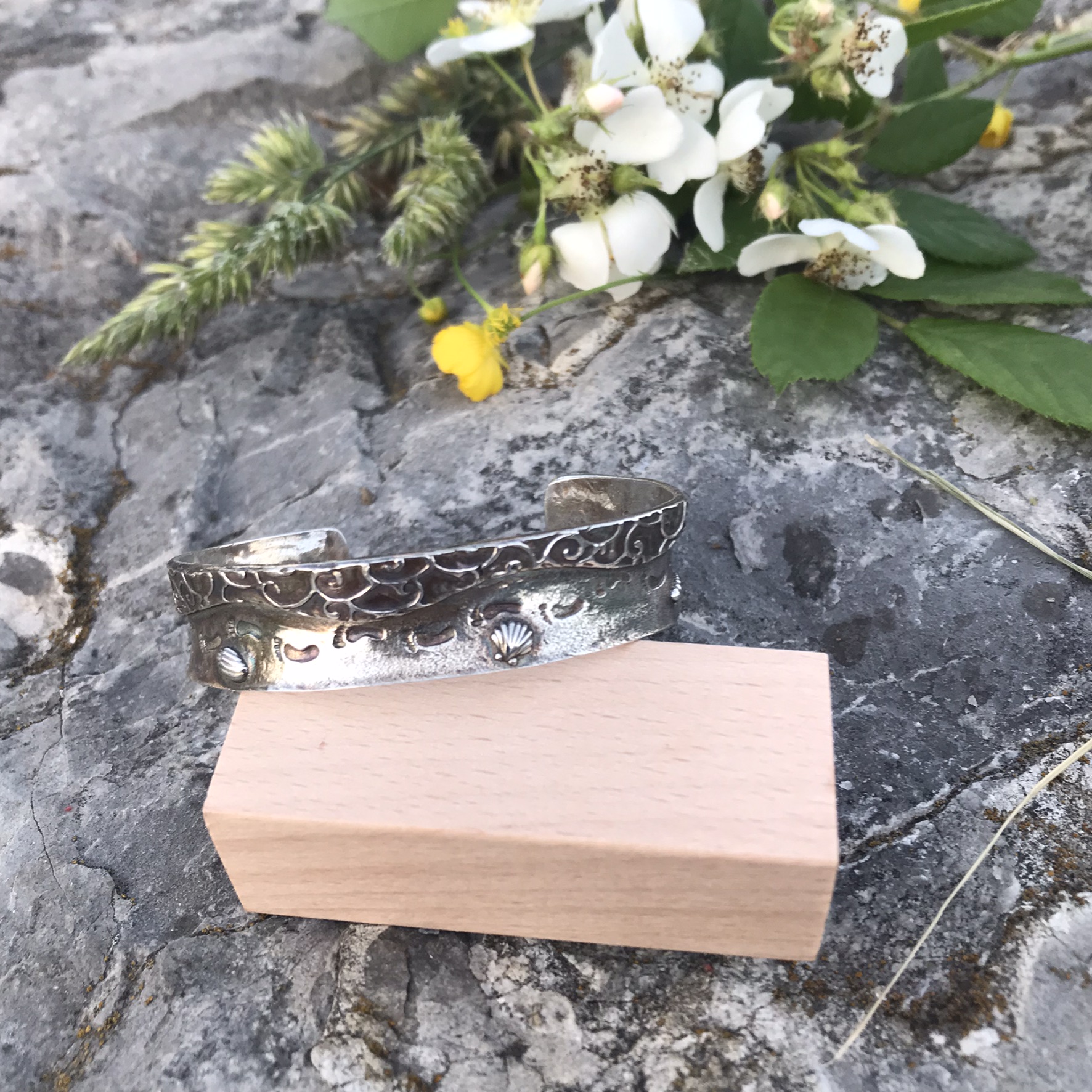 Never Alone Cuff Bracelet- 1/2 inch wide cuff bracelet created in sterling silver metal clay and showing a beach scene with water, shells and footprints in the sand propped on wooden block on rock