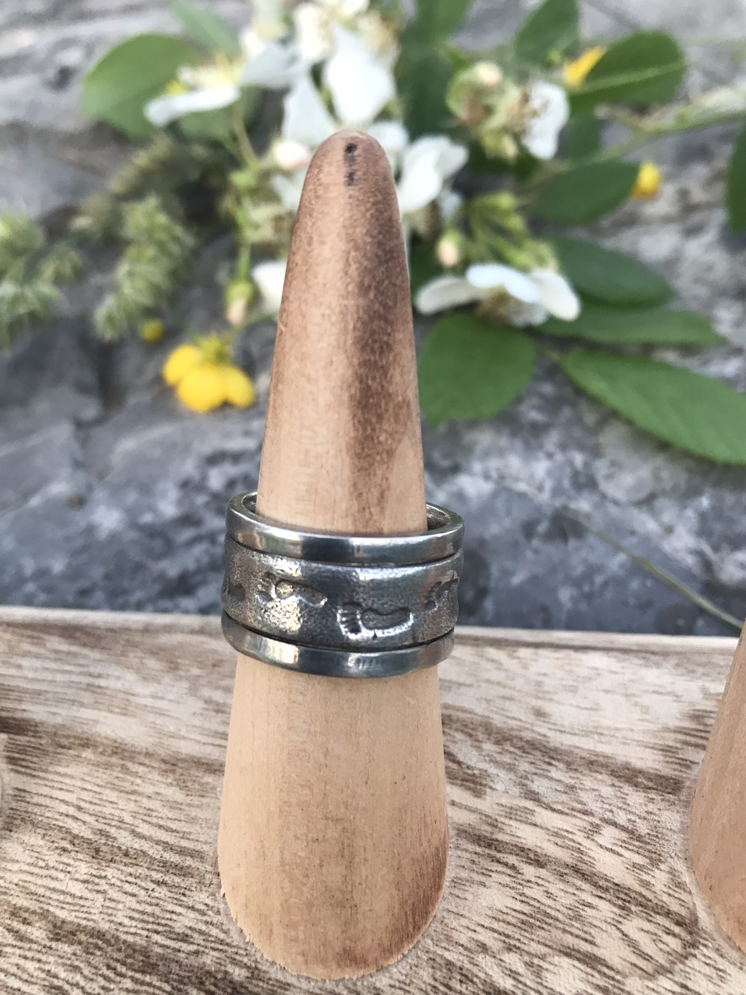 Never Alone Wide Silver Ring Band- roughly 1/2 inch wide ring created in sterling silver metal clay and showing a smooth sand texture imprinted with footprints sitting on a ring display