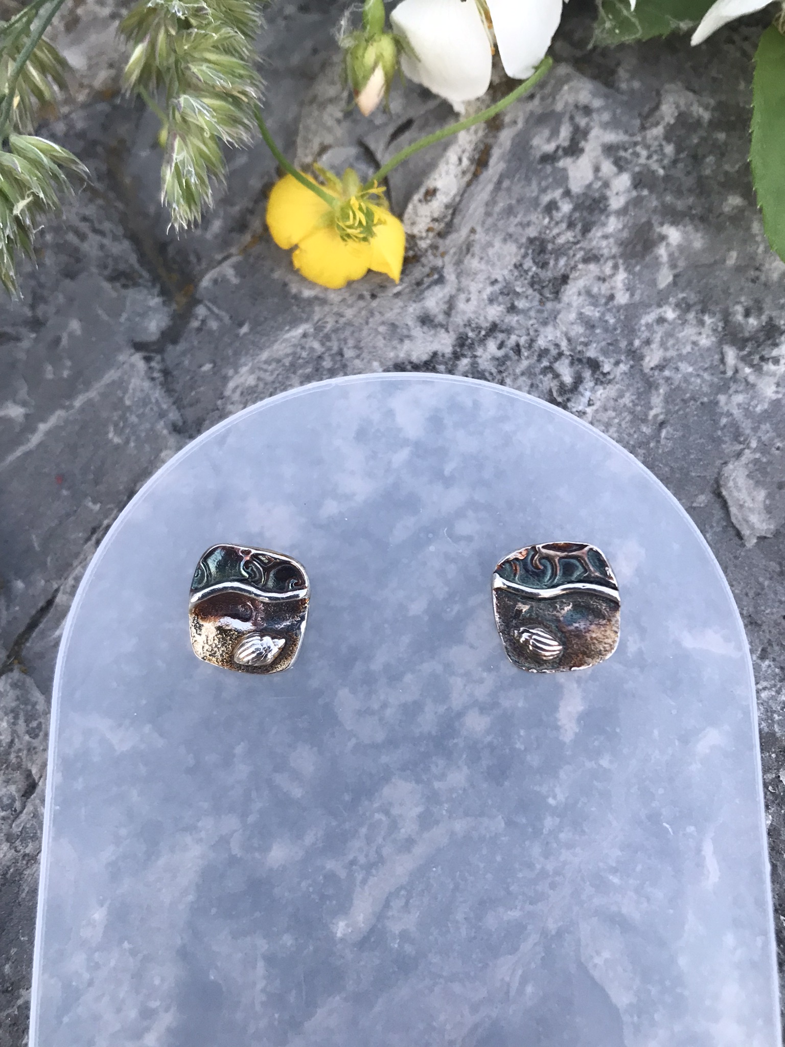 Never Alone Small Cushion Post Earrings- rounded square shaped earrings created in fine silver metal clay and showing a beach scene with water, shells and footprints in the sand with titanium posts