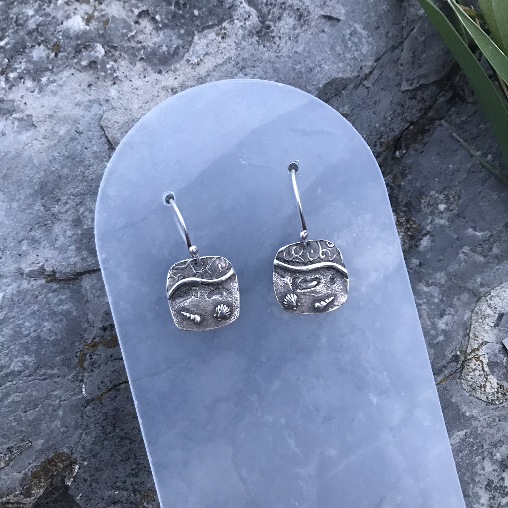 Never Alone Drop Earrings- square shaped drops created in fine silver metal clay and showing a beach scene with water, shells and footprints in the sand with handmade sterling silver earwires on earring display with rock backdrop